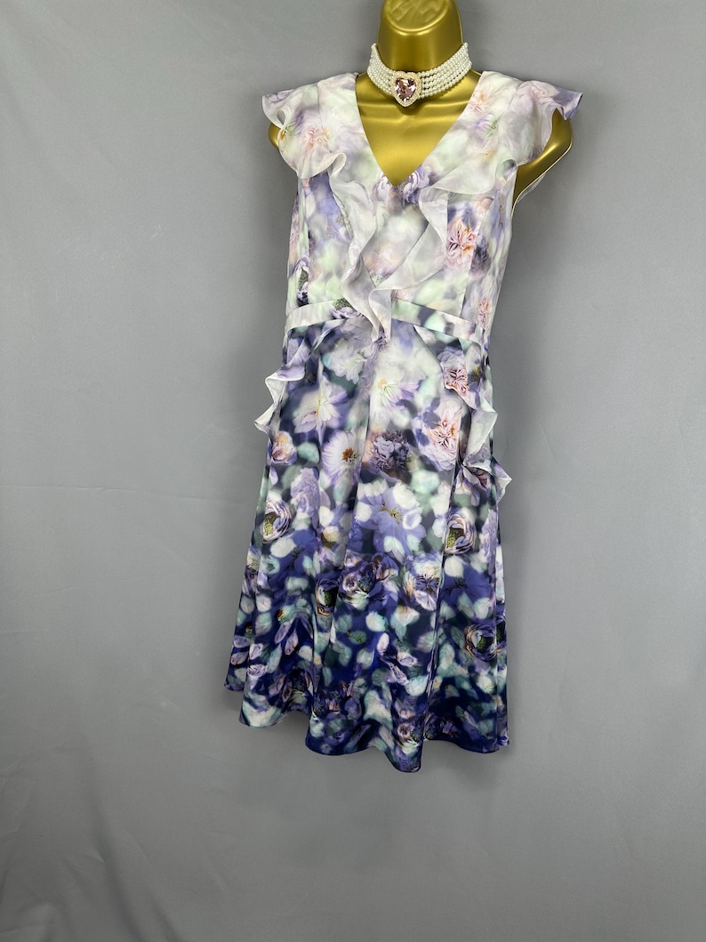 May include: A sleeveless floral print dress with a V-neck and ruffled trim. The dress features a white, purple, and blue floral pattern. A white choker necklace with a pink heart-shaped pendant is also visible.