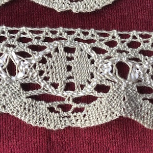 May include: A close-up of a beige crocheted lace trim with a delicate pattern. The lace has a slightly wavy edge and is made of fine threads.