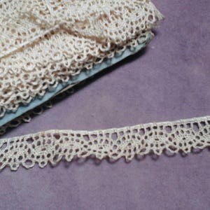 May include: A close-up of a spool of ivory-colored lace trim. The lace is delicate and intricate, with a scalloped edge. The lace is folded and rolled up on a blue spool.