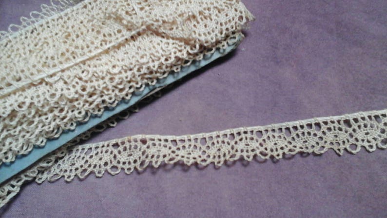 May include: A close-up of a spool of ivory-colored lace trim. The lace is delicate and intricate, with a scalloped edge. The lace is folded and rolled up on a blue spool.