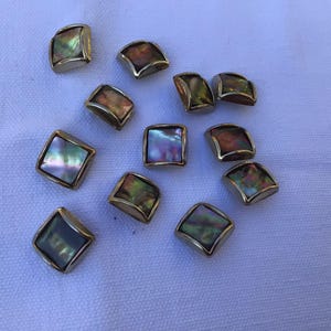 Puede incluir: Un conjunto de botones cuadrados y de bordes curvos con incrustaciones de concha de abulón iridiscente y marcos de metal dorado. Los botones están dispersos sobre una superficie blanca, mostrando sus superficies coloridas y reflectantes.