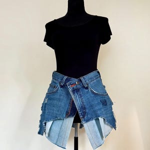 May include: A distinctive denim skirt with a deconstructed design. The skirt is made from repurposed blue jeans, featuring frayed edges and a wrap-around style. The skirt is displayed on a black mannequin, showcasing its distinctive shape and texture.