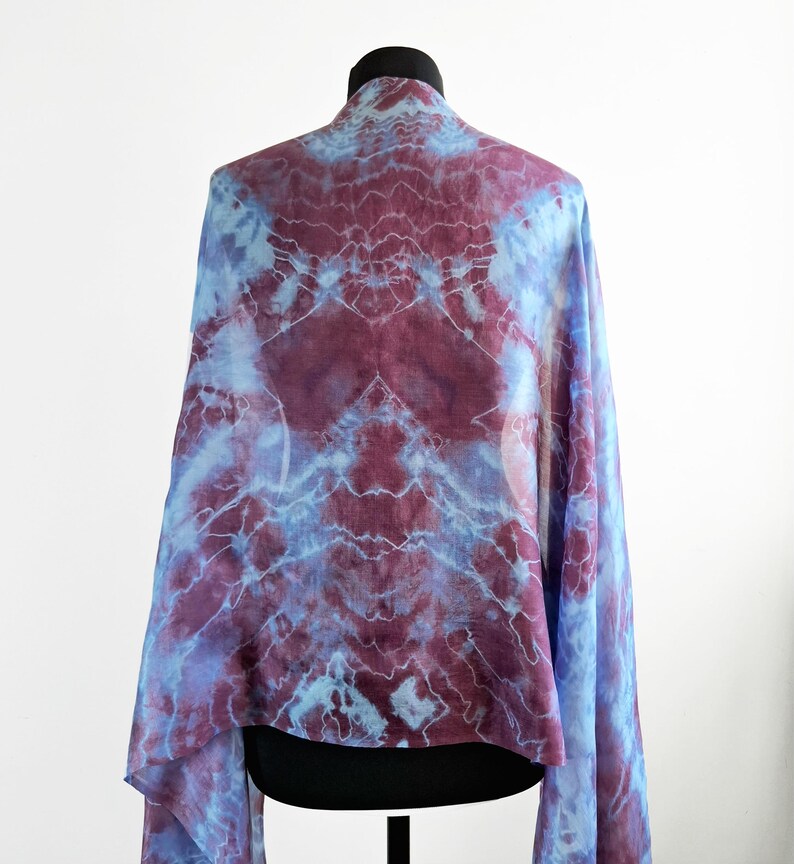A light blue and purple silk scarf with a tie dye pattern. Hand painted blue marsala silk shawl. The scarf is draped over a black mannequin.