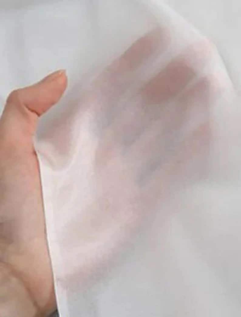 May include: Close-up of a sheer, white fabric. The fabric is held up, allowing light to pass through, revealing the hand behind it. The material appears delicate and lightweight, with a soft, flowing texture.
