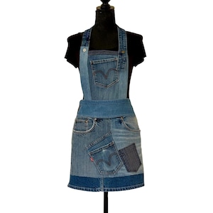 Puede incluir: Un delantal de mezclilla con diseño de retazos hecho de jeans reciclados. El delantal presenta varios tonos de mezclilla azul, incluyendo bolsillos y una cintura. El babero tiene un cierre de botón y se exhibe en un maniquí negro.