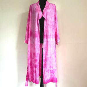 May include: A long, flowing pink robe with a tie-dye pattern, displayed on a black dress form. The robe features long sleeves and a draped, open front. The color palette includes shades of pink and white.