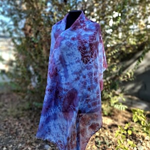 A light blue and purple silk scarf with a tie dye pattern. Hand painted blue marsala silk shawl. The scarf is draped over a black mannequin.