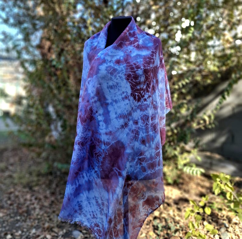 A light blue and purple silk scarf with a tie dye pattern. Hand painted blue marsala silk shawl. The scarf is draped over a black mannequin.