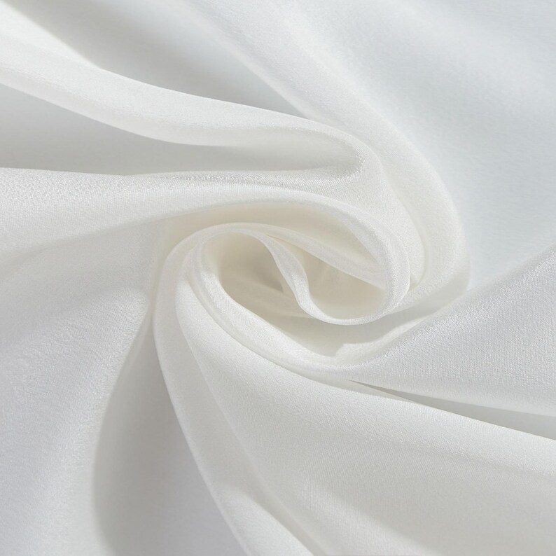 May include: Close-up of white fabric with soft folds and a silky texture. The fabric appears to be lightweight and flowing, with highlights and shadows creating depth. The overall impression is one of elegance and smoothness.