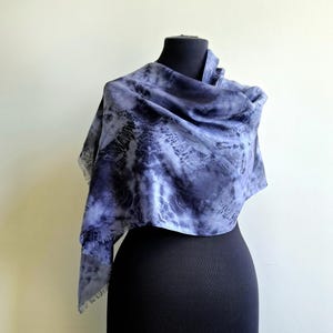 May include: A blue and grey tie-dye scarf draped over a black mannequin. The scarf's fabric displays a flowing design with intricate patterns. The edges of the scarf are slightly frayed, adding to its unique appearance.