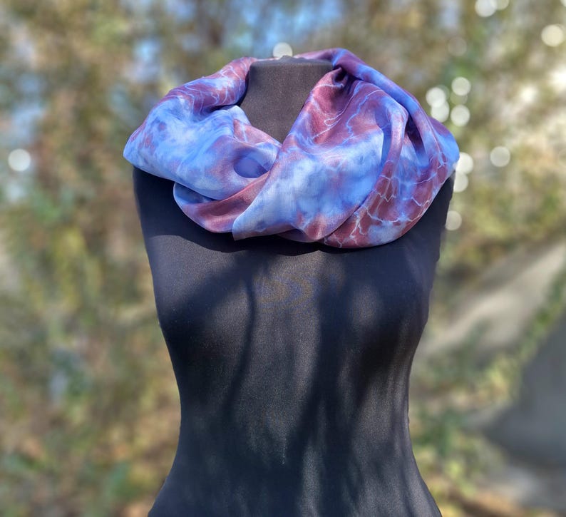 A light blue and purple silk scarf with a tie dye pattern. Hand painted blue marsala silk shawl. The scarf is draped over a black mannequin.