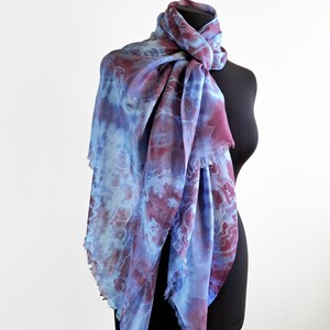 A light blue and purple silk scarf with a tie dye pattern. Hand painted blue marsala silk shawl. The scarf is draped over a black mannequin.