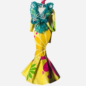 May include: A decorative Day of the Dead figurine of a skeleton in a yellow floral dress with a ruffled teal top and matching hat. The dress has pink and green accents. The skeleton has a painted face and a pink tongue.