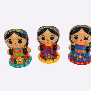 May include: Three colorful ceramic dolls with black hair, wearing brightly colored dresses with floral patterns. The dolls have smiling faces with large eyes and are a traditional Mexican folk art.