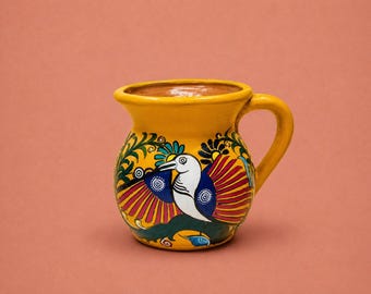 May include: A yellow ceramic mug with a rounded body and a single handle. The exterior features a hand-painted design of a white bird with blue and red wings, surrounded by green foliage and floral patterns against a bright yellow background.