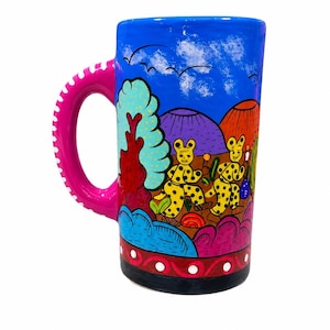 May include: A tall, hand-painted ceramic mug with a vibrant scene of yellow spotted creatures, trees, and mountains under a blue sky. The handle is bright pink with white dots. The mug has a black base and a decorative border.