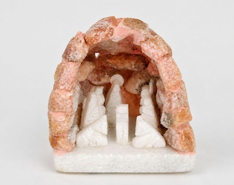 Puede incluir: Un belén de piedra tallada. La estructura tiene forma de cueva con un exterior rosa y beige. En el interior, figuras de piedra blanca representan la Sagrada Familia. La base es blanca y el diseño general es detallado y artístico.