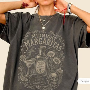 Op de afbeelding: Een antracietgrijs t-shirt met korte mouwen en een grafische print met de tekst Midnight Margaritas, een cocktailglas, een fles met het label Potion for the Perfect Margarita, een schedel, bloemen en een recept met 2 oz tequila, 1 oz triple sec, 1 oz limoensap, 1/2 oz agavenectar en 1/2 oz dash of magic.