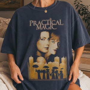 Op de afbeelding: Een donker antracietgrijs t-shirt met korte mouwen en een grafische print van de filmtitel Practical Magic. Het ontwerp bevat portretten van twee personen boven een rij brandende kaarsen en silhouetten van figuren met paraplu's.