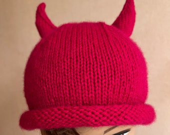 May include: A bright pink knitted hat with two horns on top. The hat is made of a soft, warm yarn and has a folded brim.