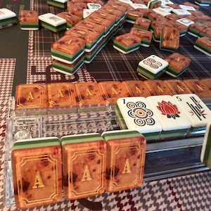 Puede incluir: Un juego de Mahjong completo con fichas naranjas, verdes y blancas. Las fichas est&aacute;n dispuestas sobre una superficie estampada. Algunas fichas tienen la letra "A" en dorado, mientras que otras presentan s&iacute;mbolos. El juego incluye un soporte de fichas de pl&aacute;stico transparente.