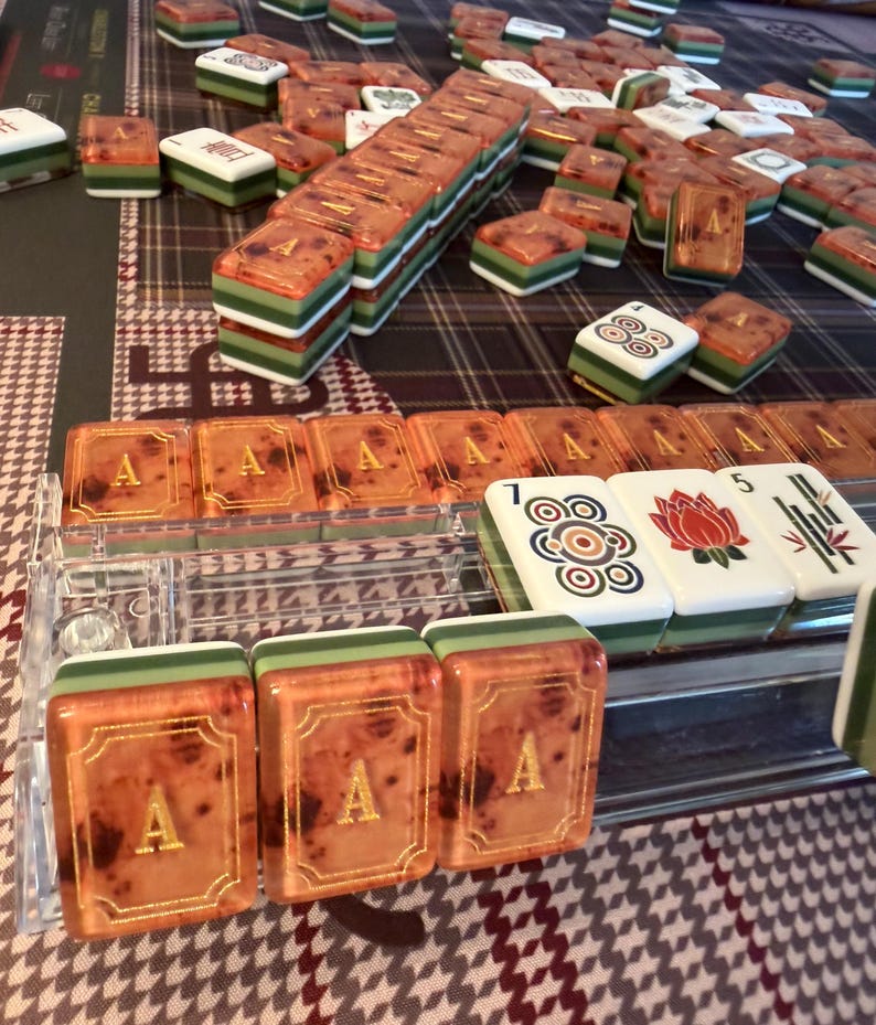 Puede incluir: Un juego de Mahjong completo con fichas naranjas, verdes y blancas. Las fichas est&aacute;n dispuestas sobre una superficie estampada. Algunas fichas tienen la letra "A" en dorado, mientras que otras presentan s&iacute;mbolos. El juego incluye un soporte de fichas de pl&aacute;stico transparente.