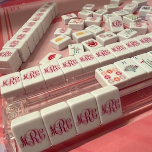 Puede incluir: Un juego de Mahjong blanco con detalles en rosa. Las fichas muestran las letras "MRC" en una escritura decorativa. Algunas fichas presentan ilustraciones coloridas. El juego est&aacute; dispuesto sobre una superficie rosa, con un soporte de pl&aacute;stico transparente.