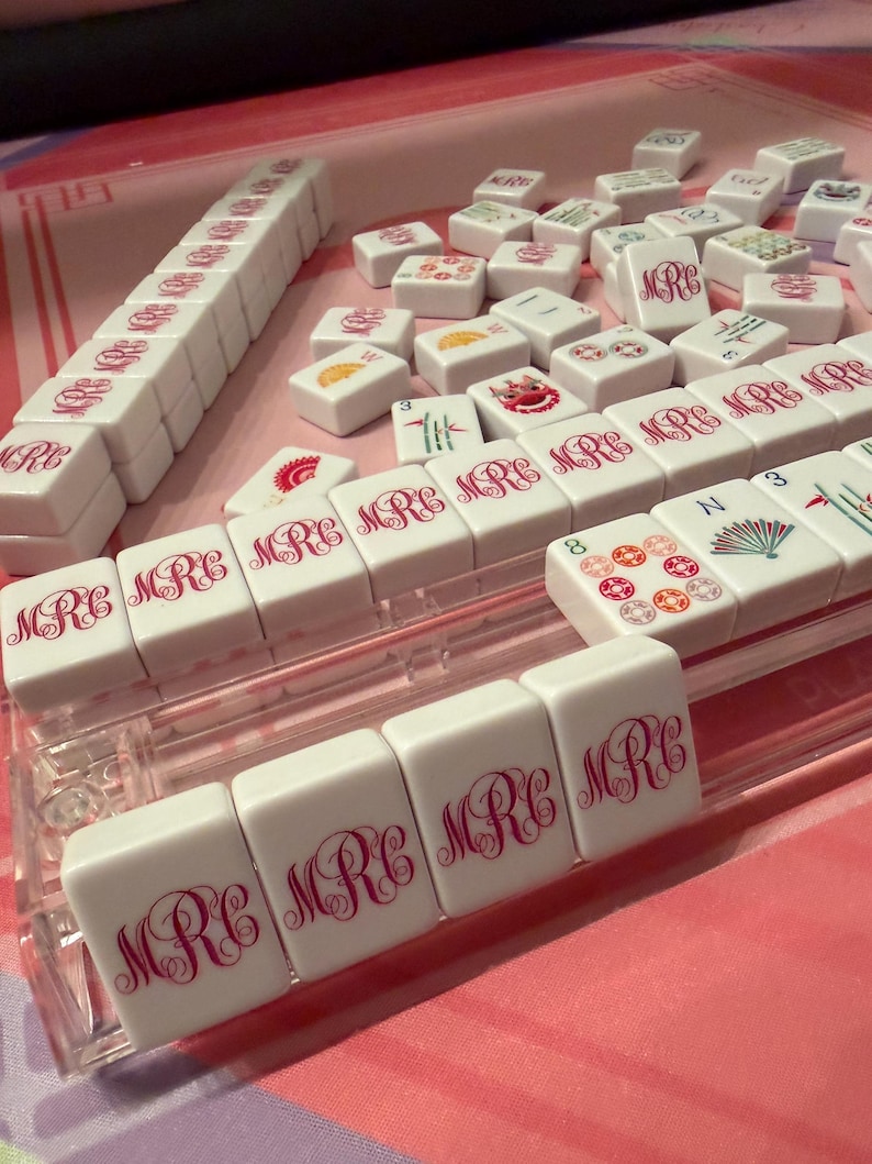 Puede incluir: Un juego de Mahjong blanco con detalles en rosa. Las fichas muestran las letras "MRC" en una escritura decorativa. Algunas fichas presentan ilustraciones coloridas. El juego est&aacute; dispuesto sobre una superficie rosa, con un soporte de pl&aacute;stico transparente.
