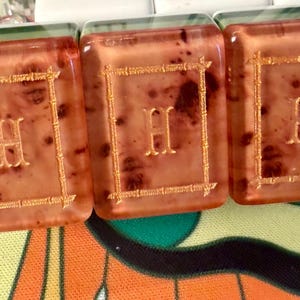 Puede incluir: Primer plano de tres fichas rectangulares transl&uacute;cidas con un tono marr&oacute;n rojizo. Cada ficha presenta la letra "H" dorada dentro de un marco dorado. Las fichas son parte de un juego, posiblemente Mahjong o similar.