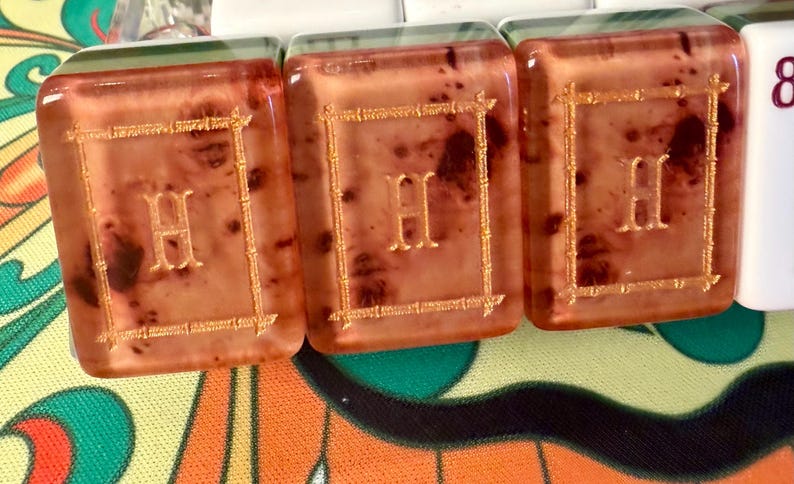 Puede incluir: Primer plano de tres fichas rectangulares transl&uacute;cidas con un tono marr&oacute;n rojizo. Cada ficha presenta la letra "H" dorada dentro de un marco dorado. Las fichas son parte de un juego, posiblemente Mahjong o similar.