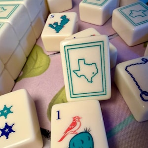 Puede incluir: Un conjunto de fichas de Mahjong blanquecinas con varios dise&ntilde;os turquesa y azul. Algunas fichas presentan un contorno de Texas, un cactus con un p&aacute;jaro y patrones de estrellas. Las fichas est&aacute;n dispuestas sobre una superficie con estampado floral.