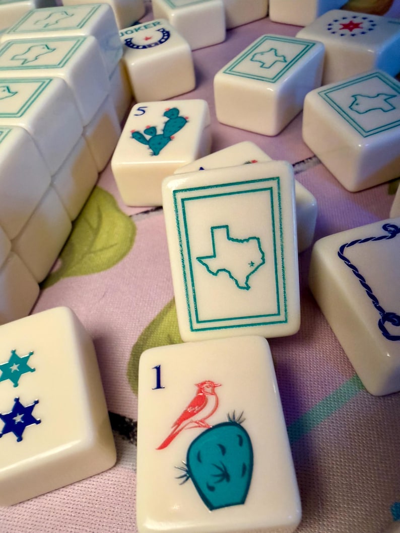 Puede incluir: Un conjunto de fichas de Mahjong blanquecinas con varios dise&ntilde;os turquesa y azul. Algunas fichas presentan un contorno de Texas, un cactus con un p&aacute;jaro y patrones de estrellas. Las fichas est&aacute;n dispuestas sobre una superficie con estampado floral.