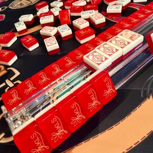 Puede incluir: Un conjunto de fichas de Mahjong rojas y blancas con varios s&iacute;mbolos y dise&ntilde;os. Las fichas est&aacute;n dispuestas sobre una superficie negra con un soporte de pl&aacute;stico transparente. Algunas fichas est&aacute;n dispersas, otras en filas. El texto "JAKLS" es visible.