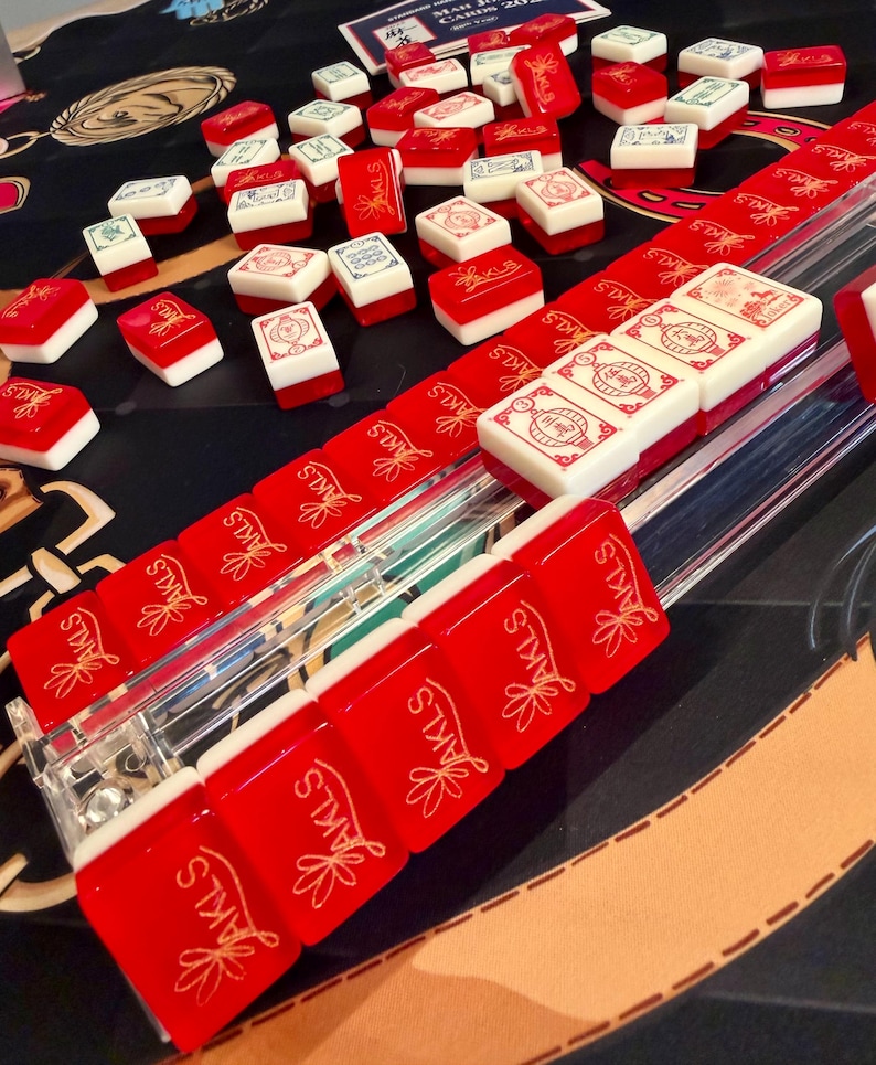 Puede incluir: Un conjunto de fichas de Mahjong rojas y blancas con varios s&iacute;mbolos y dise&ntilde;os. Las fichas est&aacute;n dispuestas sobre una superficie negra con un soporte de pl&aacute;stico transparente. Algunas fichas est&aacute;n dispersas, otras en filas. El texto "JAKLS" es visible.