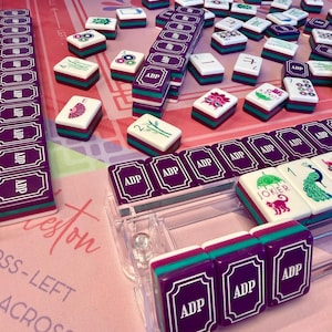Puede incluir: Un juego de Mahjong completo con fichas moradas, turquesas y blancas. Las fichas est&aacute;n dispuestas sobre una superficie rosa con texto. El juego incluye estantes y fichas con varios s&iacute;mbolos y las letras "ADP".