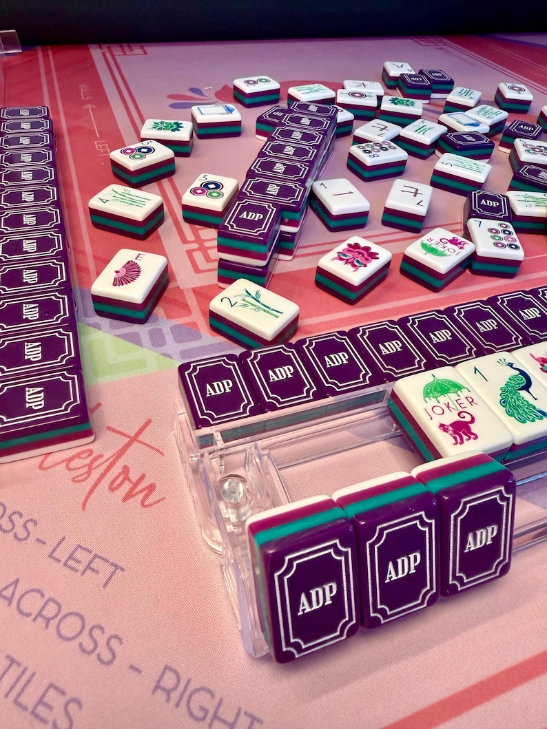 Puede incluir: Un juego de Mahjong completo con fichas moradas, turquesas y blancas. Las fichas est&aacute;n dispuestas sobre una superficie rosa con texto. El juego incluye estantes y fichas con varios s&iacute;mbolos y las letras "ADP".