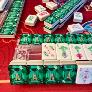 Puede incluir: Un conjunto de fichas de Mahjong con un llamativo dise&ntilde;o de piedras preciosas verdes. Las fichas est&aacute;n dispuestas sobre una superficie roja, algunas en estuches de pl&aacute;stico transparente. Varias fichas muestran s&iacute;mbolos y caracteres, incluyendo la letra "A" y la palabra "Joker".