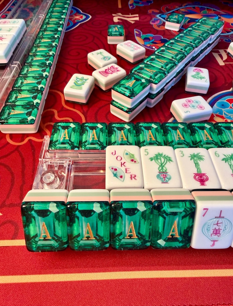 Puede incluir: Un conjunto de fichas de Mahjong con un llamativo dise&ntilde;o de piedras preciosas verdes. Las fichas est&aacute;n dispuestas sobre una superficie roja, algunas en estuches de pl&aacute;stico transparente. Varias fichas muestran s&iacute;mbolos y caracteres, incluyendo la letra "A" y la palabra "Joker".