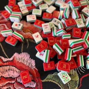 Puede incluir: Un conjunto de fichas de Mahjong rojas, blancas y verdes dispersas sobre una superficie negra con un diseño floral. Las fichas presentan varios símbolos y caracteres, algunos con las letras "PSC".