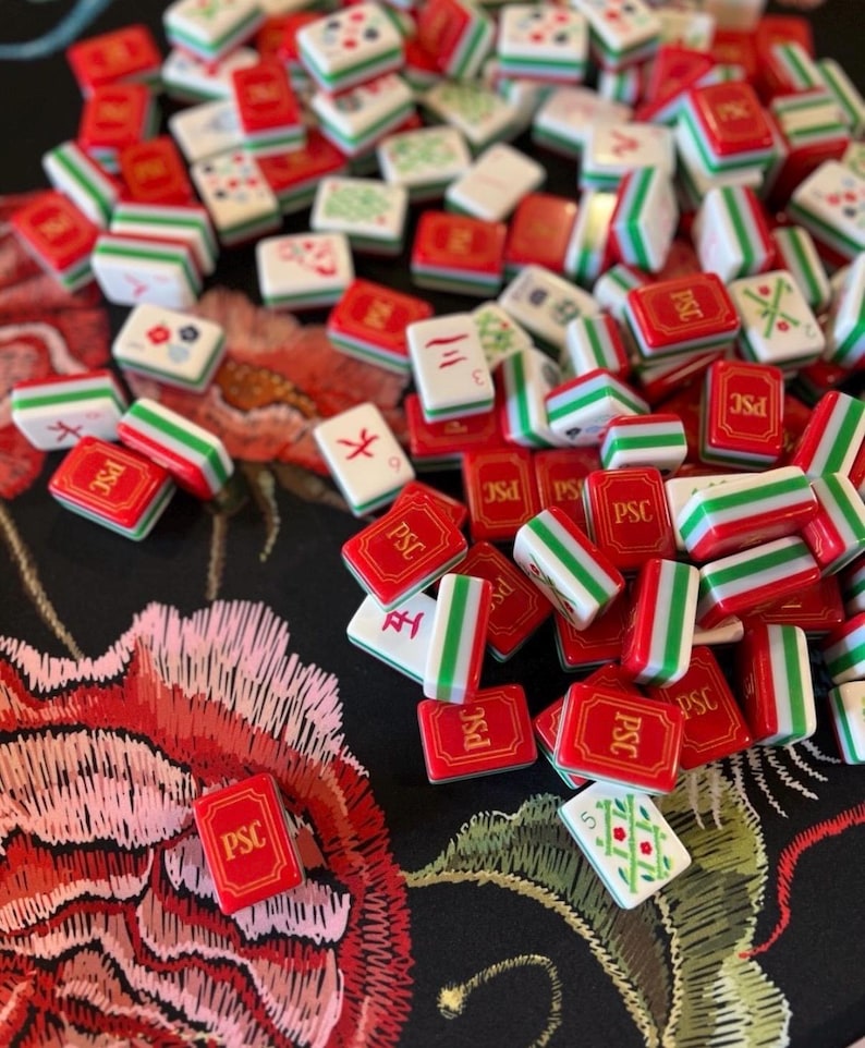Puede incluir: Un conjunto de fichas de Mahjong rojas, blancas y verdes dispersas sobre una superficie negra con un dise&ntilde;o floral. Las fichas presentan varios s&iacute;mbolos y caracteres, algunos con las letras "PSC".