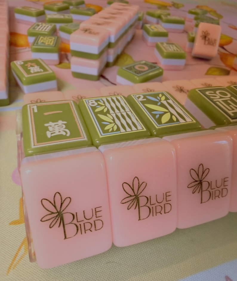 Puede incluir: Un conjunto de fichas de Mahjong en tonos rosa, verde y blanco. Algunas fichas presentan caracteres chinos y dise&ntilde;os de bamb&uacute;, mientras que otras muestran el texto "BLUE BIRD" con un gr&aacute;fico floral. Las fichas est&aacute;n dispuestas sobre una superficie estampada.