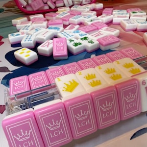 Puede incluir: Un conjunto de fichas de Mahjong rosas y blancas, algunas con el texto "LCH" y un dise&ntilde;o de corona. Las fichas est&aacute;n dispersas sobre una superficie, con algunas en una caja de pl&aacute;stico transparente y una bolsa rosa.