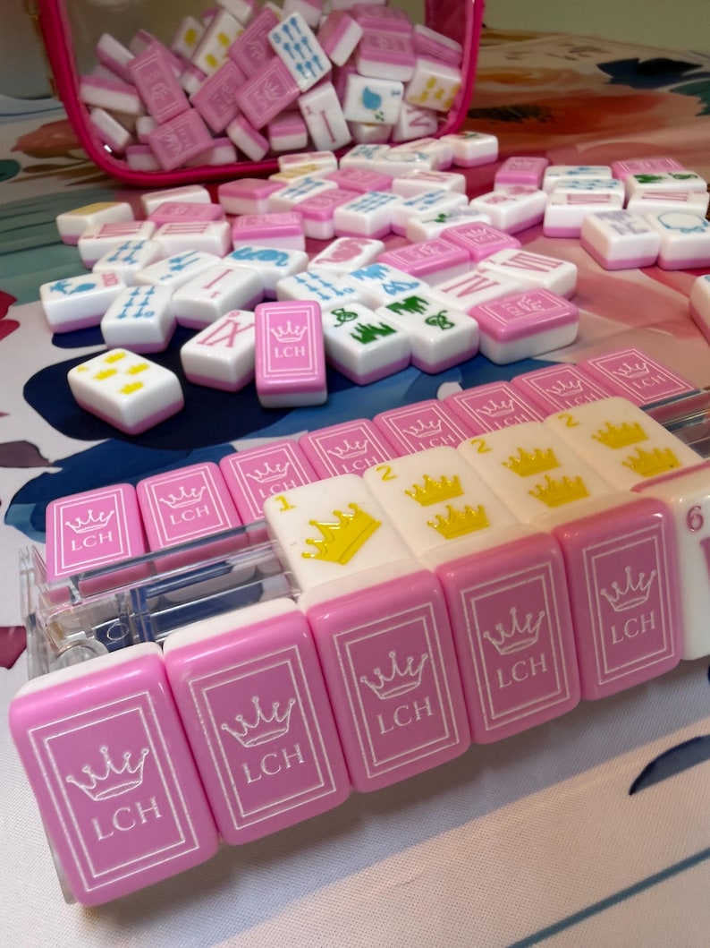 Puede incluir: Un conjunto de fichas de Mahjong rosas y blancas, algunas con el texto "LCH" y un dise&ntilde;o de corona. Las fichas est&aacute;n dispersas sobre una superficie, con algunas en una caja de pl&aacute;stico transparente y una bolsa rosa.