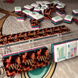 Puede incluir: Una colecci&oacute;n de coloridas fichas de Mahjong. Las fichas presentan una mezcla de patrones blancos, turquesa, rosa y carey. Algunas fichas muestran las letras "vWs" y otras caracteres chinos y n&uacute;meros. Las fichas est&aacute;n dispuestas sobre una superficie estampada.