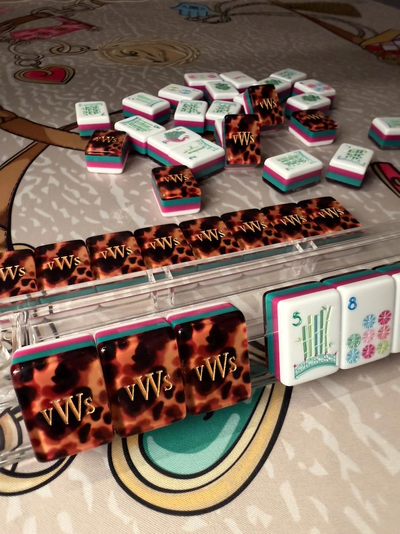 Puede incluir: Una colecci&oacute;n de coloridas fichas de Mahjong. Las fichas presentan una mezcla de patrones blancos, turquesa, rosa y carey. Algunas fichas muestran las letras "vWs" y otras caracteres chinos y n&uacute;meros. Las fichas est&aacute;n dispuestas sobre una superficie estampada.