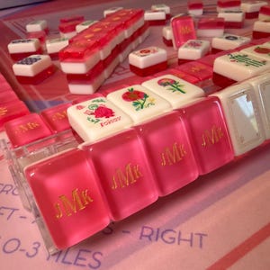 Puede incluir: Primer plano de un juego de Mahjong con fichas rosas y blancas. Algunas fichas son rosas con letras doradas, otras blancas con dise&ntilde;os florales. El juego est&aacute; dispuesto sobre una superficie rosa.