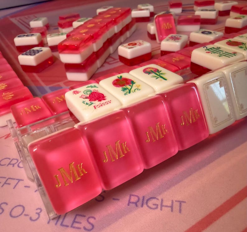Puede incluir: Primer plano de un juego de Mahjong con fichas rosas y blancas. Algunas fichas son rosas con letras doradas, otras blancas con dise&ntilde;os florales. El juego est&aacute; dispuesto sobre una superficie rosa.