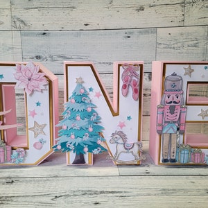 May include: Pink, white, and gold cardboard letters spelling "ONE" decorated with a ballerina, a Christmas tree, a nutcracker, and a rocking horse. The letters are decorated with gold glitter and stars.