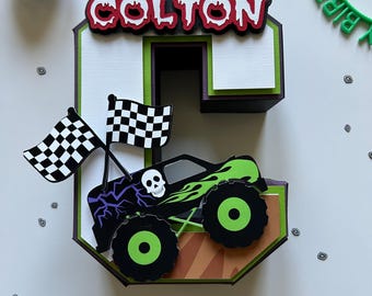May include: A layered papercraft decoration in the shape of the number 6, featuring a monster truck design. The name "COLTON" is displayed at the top. Includes checkered flags and a green "HAPPY BIRTHDAY" headband.