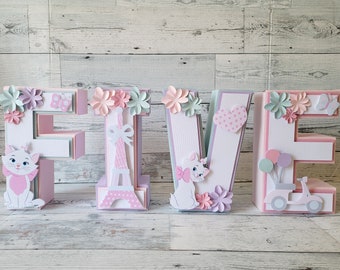 Op de afbeelding: Vijf roze, groene en witte kartonnen letters die het woord "VIJF" spellen met verschillende decoraties, waaronder bloemen, vlinders, een hart, een scooter, ballonnen en een kat met een strik.